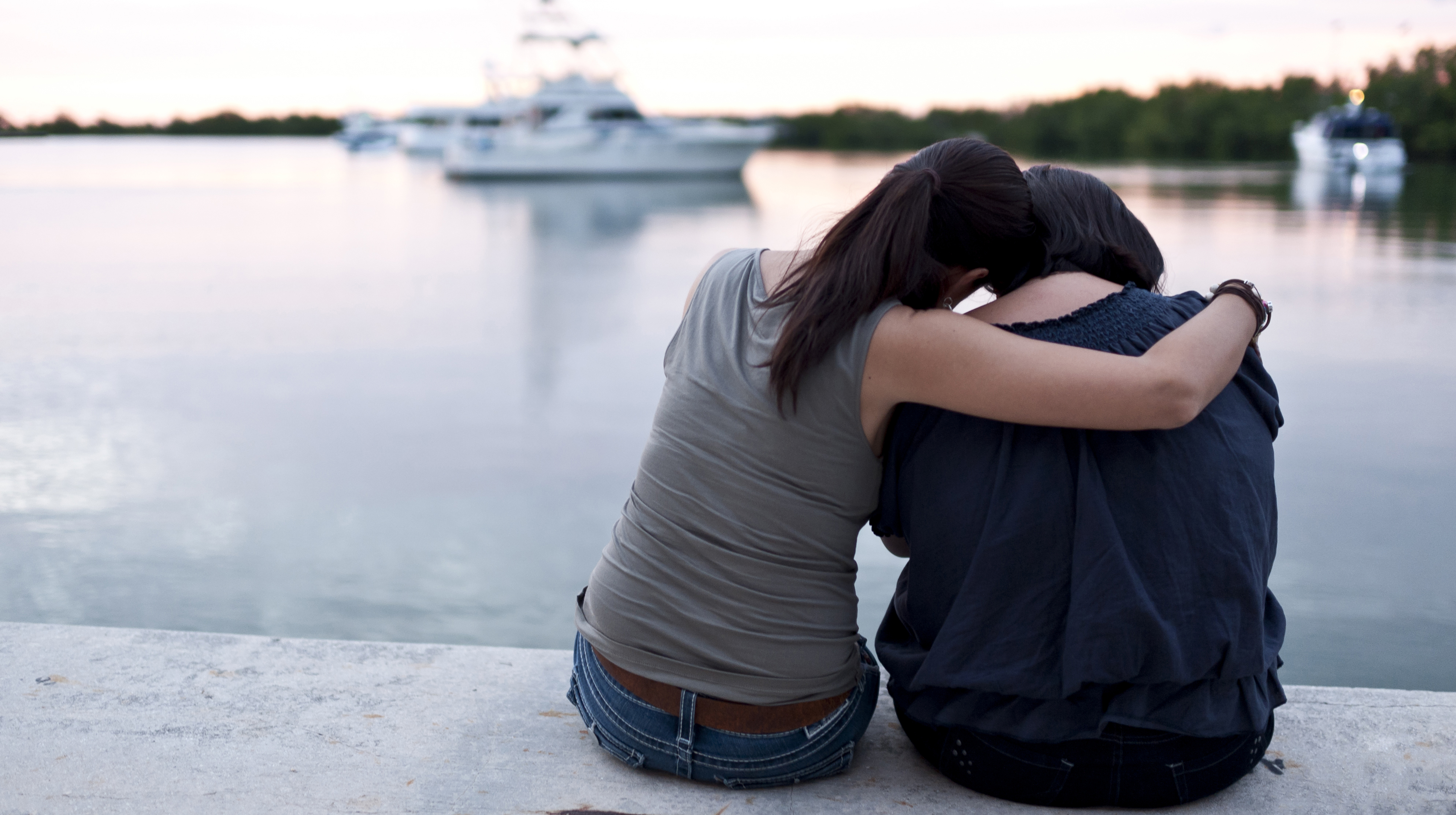 Picture, from behind, of two female students sitting by a lake; one is consoling the other with an arm around her shoulder.